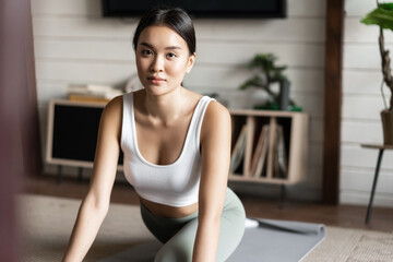 Naklejka na ściany i meble Asian girl doing yoga stretching at home, workout in her living room, wearing activewear
