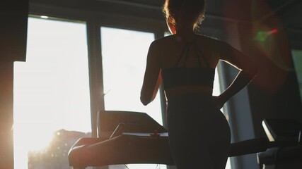 A young woman is running on a treadmill and listening to music. She's running near the window. The evening sun is shining. Training in the gym. 4K