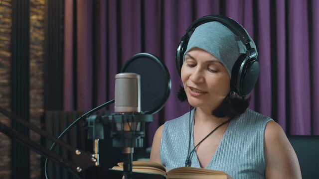 A woman in a recording studio reads aloud on a microphone an excerpt from a book. Studio recording of an audiobook. Microphone and headphones for working on audio material.