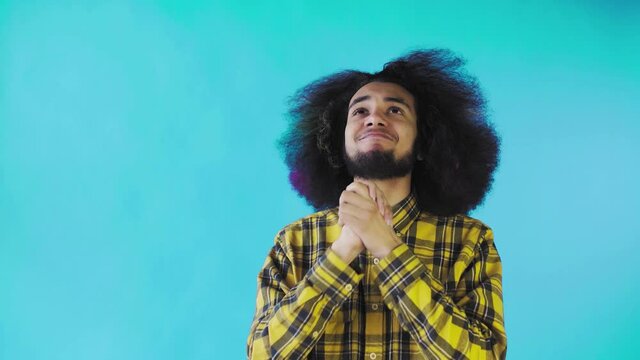 A young man with an African hairstyle on a blue background crosses his fingers and prays. Emotions on a colored background.