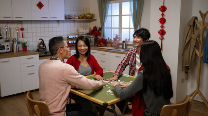 smiling asian family of four having fun with mahjong game and chatting lively in the evening at home during chinese lunar new year holiday. chinese text at background translation: luck