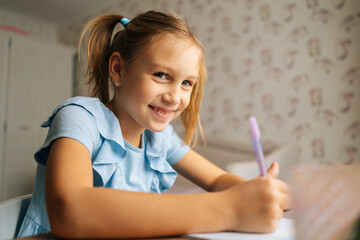 Close-up of cheerful elementary little child girl writing doing homework sitting at home table by window, looking at camera. Happy primary kid studying alone making note with pen at bedroom.