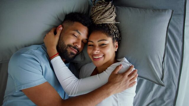 Top view of multiracial young couple in love lying on bed and cuddling together indoors at home, looking at camera.