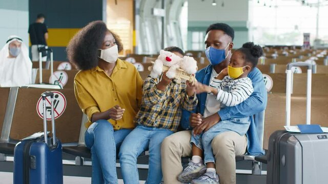 Happy African American family with two children in protective masks sitting in waiting room before departure and talking while playing with toys.