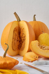 Several pumpkins on a light table, on a white background. One pumpkin is cut, pieces of pumpkin with seeds are next to it.