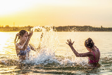 Two sisters splashing water playing in the lake at sunset background