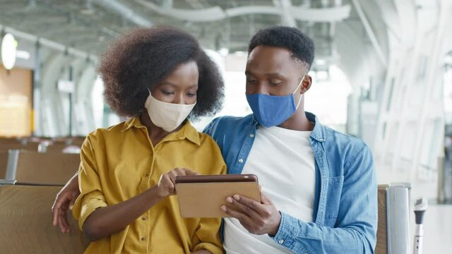 Close up of young african american couple in protective masks sitting in airport waiting room and looking at photos on tablet.