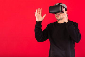 Young man with short beard, wearing black sweatshirt, cap and virtual reality glasses amazed, touching something virtually, on red background. Technology, VR, computing and hobbies concept.