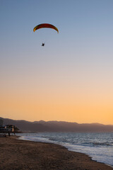 Motor hang glider flying high in the air over the beach and ocean at sunset hour. Sport and recreation concept.
