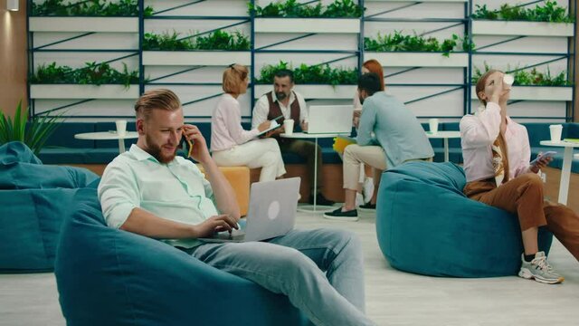 A blonde haired business man is talking on the phone while sitting on a teal colored bean bag and doing work on his laptop, other people around him are also busy. Arri Alexa Mini.