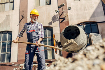 A senior construction worker is putting cement into a cement blender while standing at the...