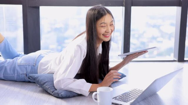 Young asian woman lying on the floor with legs up,listen watch digital class e-learning online for employee reskill upskill on laptop, tablet at home. Business school lesson education for adult people