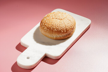 One white wheat burger bun with sesame seeds on marble board on pink seamless surface, hard light