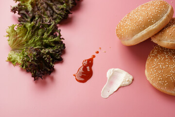 Burger ingredients: white wheat buns with sesame seeds, splashes of mayonnaise and ketchup, salad leaves on pink seamless surface