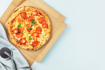 Natural italian big pepperoni pizza with thin dough, salami, mozzarella, cherry tomatoes and basil in a craft box on a blue background with copy space. Mediterranean cuisine. Pizza delivery. Flat lay