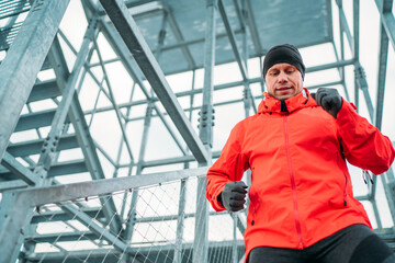 Fototapeta premium Waist portrait of smiling runner man dressed bright red softshell sporty clothes and gloves running down by huge steel industrial stairs in cold winter day. People healthy lifestyle concept photo.