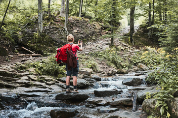 Taking pictures from vacation. Woman with backpack taking photos of landscape using smartphone camera standing on rock on mountain stream during summer trip