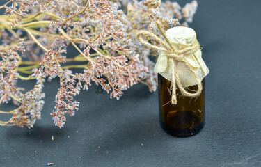 A small bottle made of dark glass with a lid made of parchment paper tied with a rope and dried herbs on a dark  background