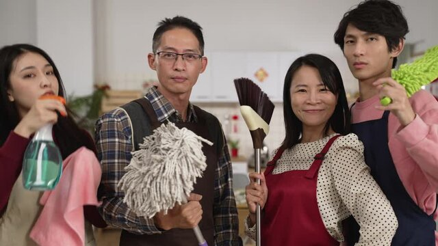 chinese father. mother. daughter and son standing up holding brooms and spray and looking at camera while posing in the kitchen at home. prepared for chinese lunar new year spring cleaning.