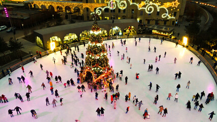 Top view of people skate on ice rink around christmas tree in park on winter holidays. Active...