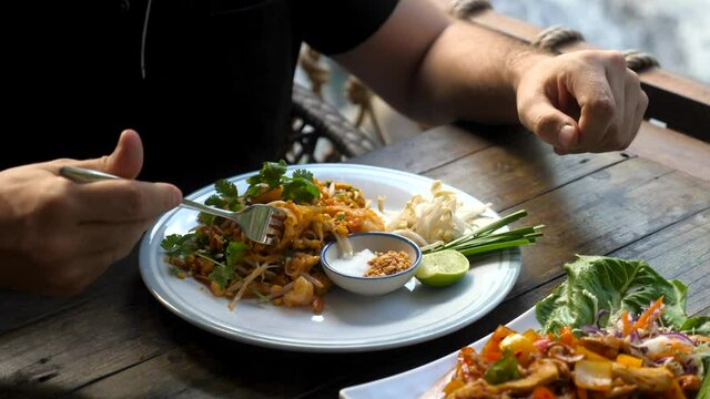Traditional thai food - pad thai. Man eating fresh food near waterfall at outdoor cafe - thai style fried rice noodle with shrimp, scrambled egg, and bean sprouts. Authentic thai local cuisine dish