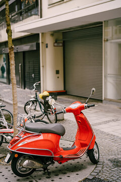 Red motor scooter stands under a tree on paving stones near the building