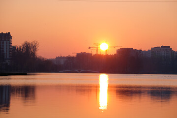 Dark silhouette of tower cranes with big setting sun at high residential apartment buildings construction site at sunset on lake shore. Real estate development