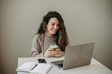 Smiling young 30 years old brunette woman with long hair uses laptop at home. He holds a mug of...