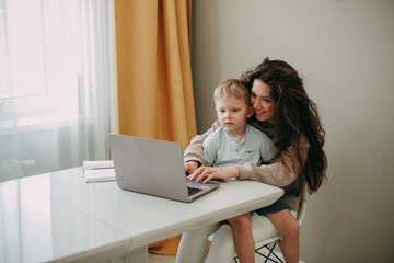 Smiling young 30 years old brunette woman with long hair sits on a chair at home. A son is sitting in her arms. Look at the laptop screen.