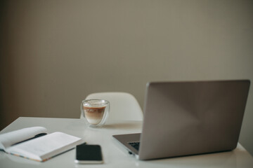 A laptop, a mug with coffee, a notebook with a pen and a mobile phone are on a white table