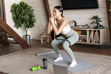 Cute asian fitness girl at home doing workout, squats with stretching elastic rope on legs, standing on floor mat, listening to online sport instructor on laptop video