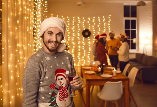Portrait of happy, smiling young man wearing traditional hat and ugly Christmas sweater with snowman holding wine glass standing against background of decorated interior at festive party with friends