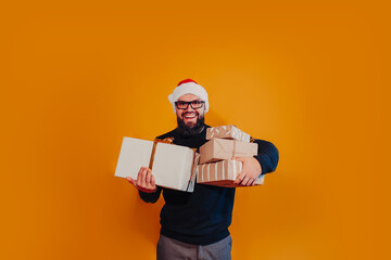 Bearded man in Santa hat holds Christmas gifts in hands. Man in Santa hat and glasses on yellow background.