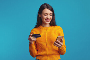 Excited pretty caucasian woman showing plastic credit card while using mobile phone isolated over blue background