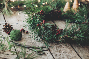 Handmade Christmas wreath with pinecones and red berries on rustic wood table, Countryside advent Christmas celebration with fir garland, candle, and Christmas lights