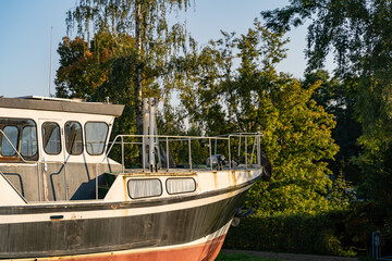 Old weathered boat in sunset sunlight. Rusty rough exterior with dirt on the surface. Wrecked ship in front of green natural trees. Traditional industry is no longer in demand.
