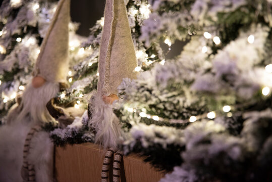 Dwa świąteczne krasnale siedzące wśród sztucznych udekorowanych ośnieżonych choinek. Ładna klimatyczna ozdoba świąteczna z białymi światełkami choinkowymi.