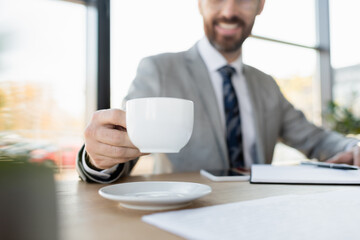 Cropped view of blurred businessman holding cup near smartphone and notebook in office.