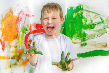 Boy with dirty hands in green paint. The child painted with his hands on the wall. Funny facial expression. Emotions, joy, carelessness, childhood. He looks at the frame and makes a grimace.