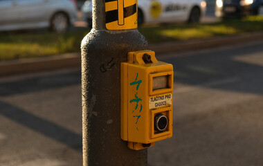 Crosswalk button for pedestrian with light warning. Defocused background with Cars drive along road and bokeh from headlights. The inscription in Czech -Tlacitko pro chodce- Button for pedestrians. 