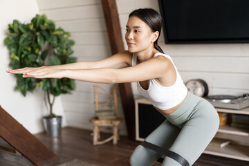Fototapeta na wymiar Cute asian fitness girl at home doing workout, squats with stretching elastic rope on her legs, exercising for fit and healthy body