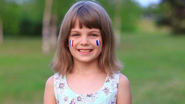 Adorable little girl smile at camera with cheeks painted in flag of France. Bastille day on July 14. The day of France. French summer holidays, travel concept.