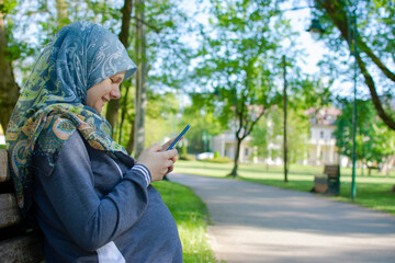Profile view of the muslim pregnant woman smiling while using mobile phone and sitting alone in the park