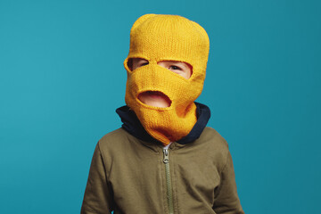 Little cute kid boy wearing yellow knitted thief mask and casual clothes, isolated against blue background