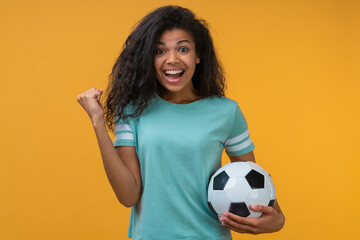 Portrait of euphoric happy football fan girl celebrating victory after betting at bookmaker's website, making winner's gesture clenching her fist while holding ball in hand - 474969158
