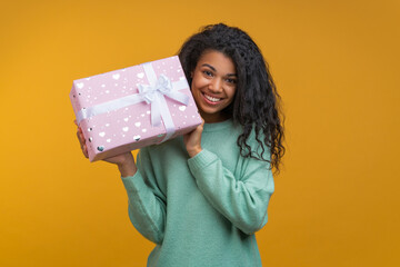 Portrait of casually dressed happy smiling girl posing with gift box wrapped in pink paper with hearts and decorated wit satin ribbon with a bow, isolated over orange yellow background