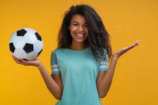 Attactive smiling girl soccer player or supporter is posing with a ball in hands isolated over bright colored yellow background