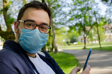 Young white man with glasses wearing face mask looking to the camera while holding mobile phone