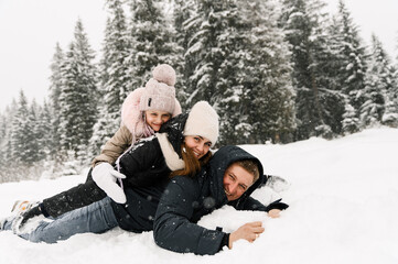 Fototapeta na wymiar Happy family have fun in winter forest. Mother, father and dauther lie on the ground and playing with snow. Family concept. Enjoying spending time together