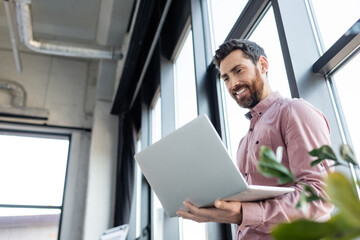 Low angle view of cheerful businessman using laptop near blurred plant in office.